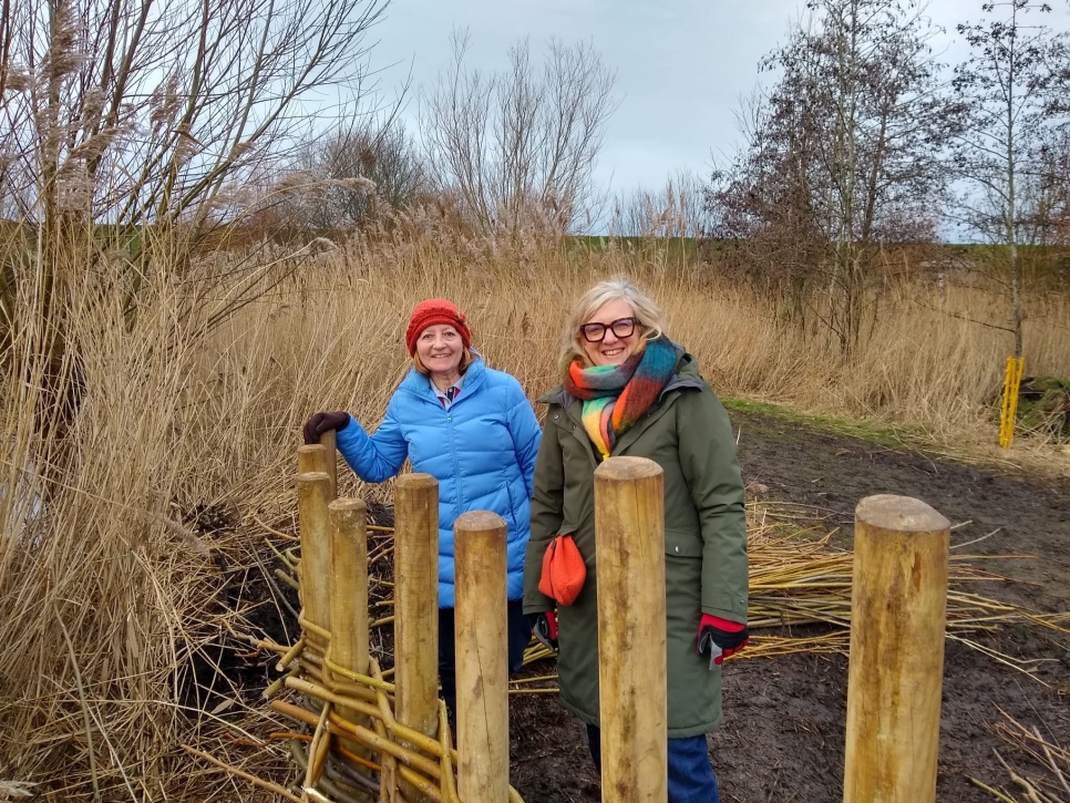 Two ladies stood next to a line of wooden fence posts surrounded by reeds.